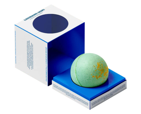 Close-up of a green bath bomb placed in a blue base with a square eco-friendly white packaging box featuring a round window opening.